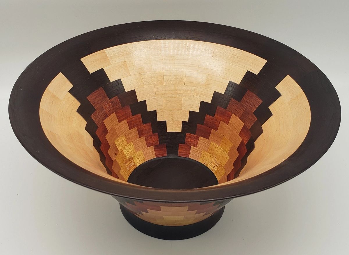 A flared segmented bowl with a stepped starburst pattern radiating from the base in pale maple, warm reddish browns and near black